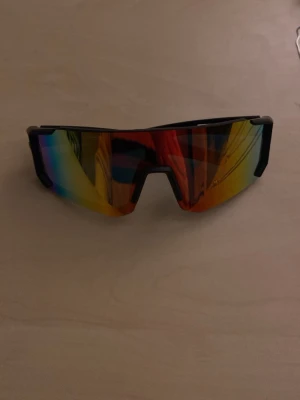 Sportiga multifärgade solglasögon - Säljer ett par sportiga solglasögon med svart båge och spegelglas i regnbågsfärger. Glasen har en kantig, futuristisk form och ger en cool vibe till din outfit. Perfekta för dig som vill sticka ut och skydda ögonen mot solen.