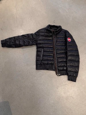 Svart Colmar dunjacka i storlek M – fint skick - Snygg svart pufferjacka från Colmar med quiltad design och hög krage. Jackan har två dragkedjeförsedda fickor och ett tydligt Colmar-märke på ärmen. Tillverkad i glansigt material som ger en modern look och håller dig varm under kalla dagar.