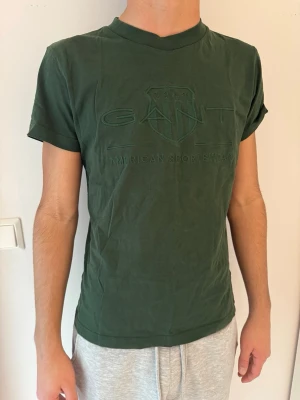 Mörkgrön GANT t-shirt - Snygg mörkgrön t-shirt från GANT med diskret logga och texten 'American Sportswear' på bröstet. Klassisk rund halsringning och korta ärmar. Tillverkad i mjuk bomull för skön känsla och enkel stil. Använd fåtal gånger