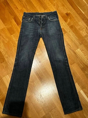 Mörkblå jeans från Nudie Jeans - Säljer ett par mörkblå jeans från Nudie Jeans med klassisk femficksmodell och kontrastsömmar. Jeansen har raka ben och snygga slitningar framtill för en avslappnad look. Perfekta till vardags och passar till det mesta.31/36