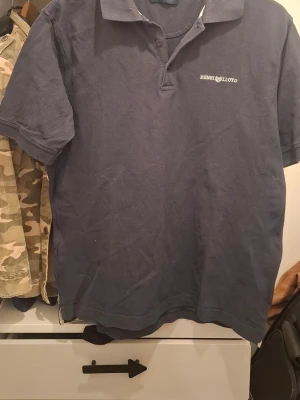 Marinblå pikétröja Henri Lloyd - Klassisk marinblå pikétröja från Henri Lloyd med korta ärmar och krage. Tröjan har diskret logga broderad på bröstet och är tillverkad i bomull för en bekväm känsla. Perfekt för en avslappnad och stilren look.