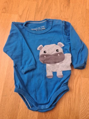 Blå body med flodhästmotiv - Supersöt blå långärmad body från petite mini i storlek 68 med ett stort grått flodhästmotiv framtill. Praktiska tryckknappar vid axeln och i grenen för enkel påklädning. Mjuk och skön bomull som passar perfekt till de minsta.