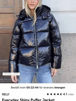 Glansig svart pufferjacka från Nelly - Snygg svart pufferjacka med glansig finish från Nelly. Jackan har en hög krage, avtagbar huva och dragkedja framtill. 