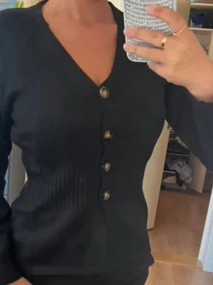 Svart ribbad kofta med knappar - Snygg svart kofta med ribbad midja och fyra dekorativa knappar framtill. Koftan har v-ringning och långa ärmar, perfekt att styla till jeans eller kjol. Materialet är stickat och ger en skön passform.