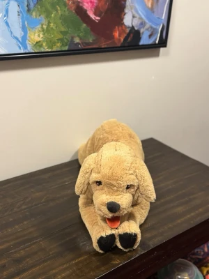 Mjukisdjur hund beige - Söt mjukishund i beige plysch med svarta tassar, svart nos och öppen mun med röd tunga. Hunden ligger ner och har mjuka, hängande öron. Perfekt som gosedjur eller dekoration i rummet.