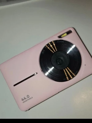 Rosa digitalkamera 44MP 16x zoom - Säljer en kompakt rosa digitalkamera med 44 megapixlar och 16x optisk zoom. Kameran har en stilren, rektangulär design med inbyggd blixt och tydliga knappar. Perfekt för dig som vill ha en smidig kamera att ta med överallt. 32GB