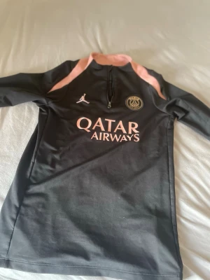 Svart PSG träningströja Jordan - Svart långärmad träningströja från PSG x Jordan med rosa detaljer och halv dragkedja. PSG-logga och Qatar Airways-tryck på bröstet, samt texten PARIS på ärmen. Perfekt för fotbollsträning eller chill. Materialet är troligtvis polyester. (äkta)