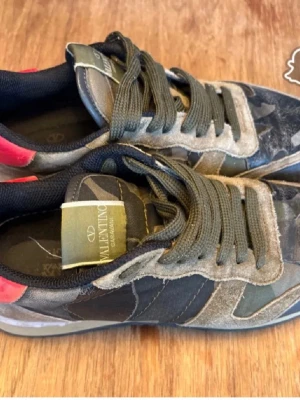 Valentino rockrunners camo sneakers med mockadetaljer - Snygga sneakers från Valentino med kamouflagemönster i grönt och grått, detaljer i mocka och läder samt en röd detalj på hälen. Skorna har gröna skosnören och låg profil. Perfekta för dig som vill sticka ut med exklusiva material och cool design.