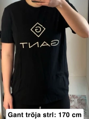 Svart Gant t-shirt med logga - Svart t-shirt från Gant med klassisk vit logga tryckt framtill. T-shirten har rund halsringning och korta ärmar. Tillverkad i mjuk bomull som känns skön mot huden. Fint skick förutom skrynklig då den inte används men går att fixa me en tvätt eller strykning