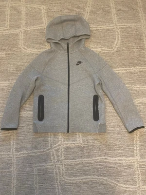 Grå Nike Tech hoddie - Snygg grå NikeTech . Hoodien har två svarta dragkedjeförsedda fickor och en klassisk huva. Den är fin med inga fläckar eller hål bara lite sliten vid ena armen annars fint skick. Vill du ha mer bilder skriv gärna ;)