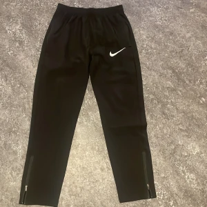 Svarta träningsbyxor från Nike, Nike miller - Svarta träningsbyxor från Nike med vit logga på låret. Byxorna har dragkedjor vid bensluten och resår i midjan. Perfekta för träning eller sportaktiviteter. Materialet är lätt och andas bra. Högra drak kedjan är sönder