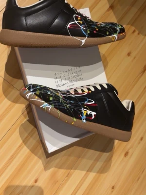 Maison Margiela svarta sneakers - Maison Margiela sneakers i svart läder med klassisk gummisula i beige. Diskret vit söm på hälen och stilren design. Perfekta för dig som gillar minimalistisk och trendig stil. Märkets logga och storlek finns på sulan.