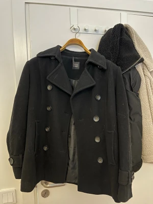 Svart skepparkavaj från ZARA - Klassisk svart skepparkavaj från ZARA med dubbelknäppning och bred krage. Jackan har två framfickor, dekorativa knappar vid ärmsluten och är fodrad för extra komfort. Perfekt för dig som gillar tidlös och stilren look.