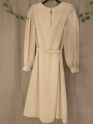 Beige långklänning med bälte - Elegant beige långklänning med rund halsringning, långa puffärmar och ett matchande bälte i midjan. Klänningen har en mjuk, flytande form och knappdetalj bak i nacken. Perfekt för dig som gillar stilrena och klassiska plagg.