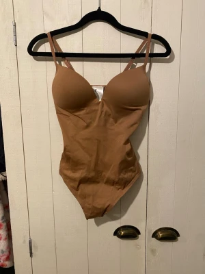 Brun body från H&M med vadderade kupor - Snygg oanvänd brun body från H&M med vadderade kupor och justerbara axelband. Tillverkad i en mjuk blandning av bomull och polyester för skön passform. Perfekt att styla under skjortor eller tröjor för en clean look.