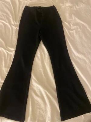 Svarta utsvängda kostymbyxor - Säljer ett par svarta kostymbyxor med utsvängda ben. Byxorna har hög midja och klassisk passform utan synliga fickor eller detaljer. Perfekta för en clean och stilren look. Materialet känns mjukt och följsamt.