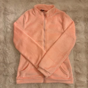 Ljusrosa fleecejacka med dragkedja - Mysig rosa/peach fleece med hel dragkedja framtill och två fickor. Den har hög krage och långa ärmar, perfekt för kyliga dagar. Materialet är mjukt och fluffigt för extra komfort. Fleecen är i storlek M men är liten i storleken. Skulle säga att den passar S/XS bättre.