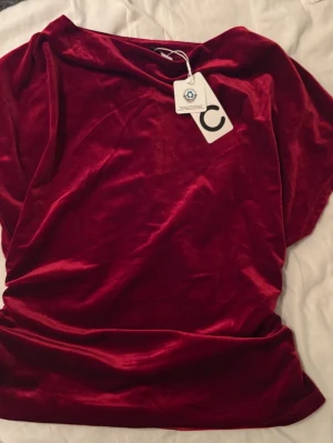 Vinröd sammetsblus med bred hals - Aldrig använd. Snygg vinröd blus i mjuk sammet med bred, något båtringad hals. Toppen har en kroppad passform och korta ärmar. Perfekt för dig som vill ha en trendig och lyxig look.