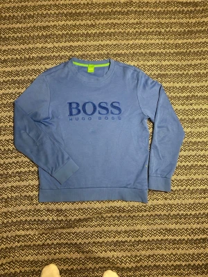 Blå sweatshirt från Hugo Boss - Snygg blå sweatshirt från Hugo Boss med broderad logga på bröstet. Tröjan har rund halsringning, ribbade muddar och en grön detalj på insidan av kragen. Slim fit-modell som passar perfekt till jeans eller chinos. Ny pris ca 1200kr