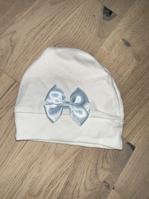 Vit mössa med ljusblå rosett - Söt vit mössa i mjukt tyg med en glansig ljusblå rosett framtill. Enkel och stilren design som passar perfekt till vardagslooken. Mössan har en uppvikt kant och är gjord i stretchigt material för skön passform.