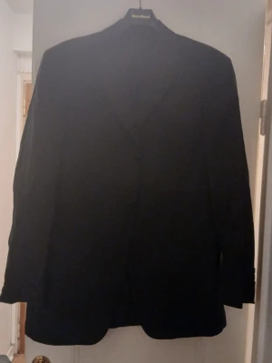 Svart klassisk kavaj - En svart kavaj med enkel knäppning och klassisk passform. Perfekt för en stilren look. Kavajen har långa ärmar och är helfärgad utan synliga detaljer. Passar till många olika outfits.