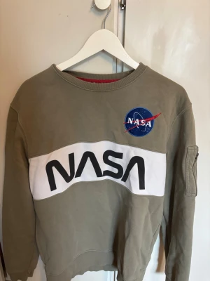 Olivgrön NASA sweatshirt - Cool olivgrön sweatshirt med stort NASA-tryck över bröstet och klassisk NASA-logga på bröstet. Tröjan har en vit panel framtill och en praktisk ficka med dragkedja på ärmen. Tillverkad i mjuk bomullsblandning och har ribbade muddar.