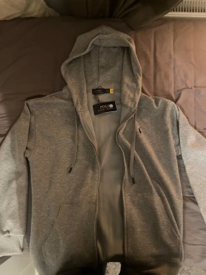 Grå hoodie från Polo Ralph Lauren - Snygg grå hoodie från Polo Ralph Lauren med dragkedja, huva med dragsko och två fickor framtill. Klassisk liten logga broderad på bröstet. Tillverkad i mjukt bomullsmaterial som är skönt mot huden. Perfekt för en avslappnad och stilren look. Passar M