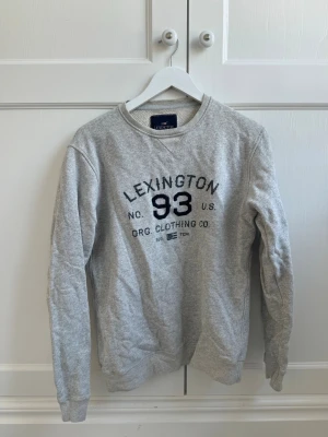 Grå sweatshirt från Lexington - Snygg grå sweatshirt från Lexington med tryck 'LEXINGTON 93' i mörkblått framtill. Klassisk rund halsringning och ribbade muddar vid ärmslut och nederkant. Tillverkad i mjukt bomullsmaterial som är skönt att bära. Perfekt för en avslappnad och sportig stil.