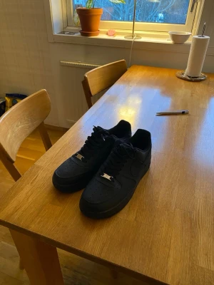 Svarta Nike Air Force 1  - Klassiska helsvarta Nike Air Force 1 sneakers i läder med perforerad tåbox och svarta skosnören. Snygg metallbricka med 'AF1' på snörningen och diskret Nike Swoosh på sidan. Perfekta för dig som gillar stilrena och tidlösa sneakers.