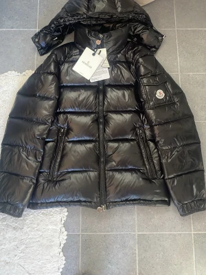 Svart dunjacka från Moncler - Snygg svart dunjacka från Moncler med glansig finish och klassisk puffer-design. Jackan har två dragkedjefickor fram, avtagbar huva och Moncler-logga på ärmen. Perfekt för kalla vinterdagar och riktigt trendig look. Storlek 2 passar M men även S 