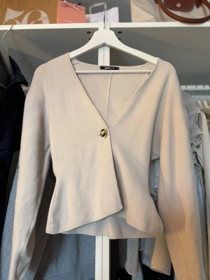 Beige kofta från Gina Tricot - mycket bra skick - Stilren beige kofta från Gina Tricot med v-ringning och en dekorativ knapp framtill. Koftan har lång ärm och en lätt utsvängd nederkant som ger en snygg siluett. 