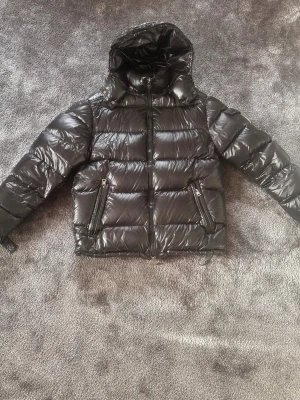 Svart dunjacka med huva från Moncler - Snygg svart dunjacka från Moncler med glansig finish och stor huva. Jackan har två dragkedjeförsedda fickor framtill och quiltad design. Perfekt för kalla vinterdagar och riktigt trendig look.