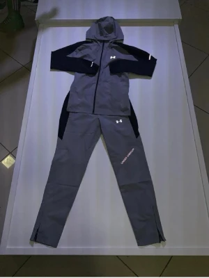 Grå träningsset från Under Armour - Snyggt grått träningsset från Under Armour med svarta detaljer. Setet består av en zip-hoodie med huva och svarta ärmar samt matchande byxor med logga på låret. Perfekt för träning eller chill. Materialet är troligtvis polyester för bra komfort och rörelsefrihet. Storlek M passar S