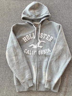 Grå Hollister hoodie med dragkedja storlek L - Grå Hollister hoodie med dragkedja och vit broderad text. Inga defekter. Storlek XL men sitter ungefär som L men kolla mått noggrant: axelbredd: 48 cm längd: 71 cm