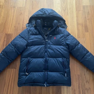 Mörkblå dunjacka från Polo Ralph Lauren - Snygg mörkblå dunjacka från  Ralph Lauren med huva och dragkedja framtill. Jackan har två fickor med dragkedja och har en innerficka och ett rött broderat logotyp på bröstet. Perfekt för kalla vinterdagar och har en klassisk, sportig look.