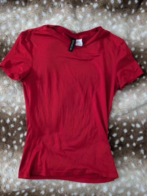 Röd t-shirt från Divided H&M - En enkel och stilren röd t-shirt från Divided H&M. Modellen är kortärmad med rund halsringning och har en figurnära passform. Perfekt att matcha med jeans eller kjol för en clean look.