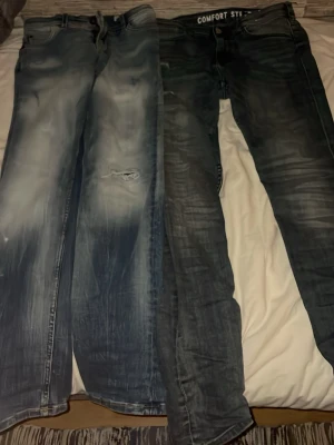 Två par blå jeans med slitningar - Säljer två par blå jeans med coola slitningar och tvättade detaljer. Ena paret har en ljusare blå ton med slitna partier och hål, medan det andra är mörkare med diskreta slitningar. Båda har klassisk femficksdesign och knappgylf. Perfekta för en avslappnad streetstil.