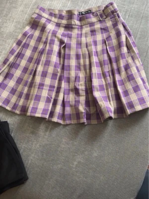 Lila rutig kortkjol från FASSTER - Säljer en superfin lila och beige rutig kortkjol från FASSTER. Kjolen har plisserade veck och ett snyggt bälte i midjan. Perfekt för dig som gillar preppy stil och vill sticka ut lite extra.