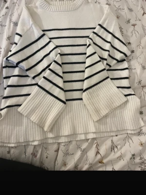 Vit stickad tröja med marinblå ränder - Säljer en vit stickad tröja med svart horisontella ränder. Tröjan har breda, ribbstickade muddar och en avslappnad passform. Perfekt för dig som gillar klassisk och stilren design.