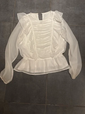 Vit volangblus med långa ärmar - Säljer en vit blus med volangdetaljer och rynk framtill. Blusen har långa, transparenta ärmar och en lätt utsvängd midja. Materialet är tunt och luftig. Knäppning i nacken med liten öppning.