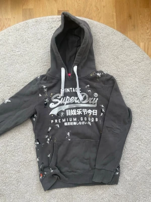 Mörkgrå hoodie från Superdry - Snygg mörkgrå hoodie från Superdry med vit logga och japansk text på bröstet. Hoodien har vit dragsko i huvan, känguruficka och ribbade muddar. Tillverkad i mjukt bomullsmaterial och har detaljer i vitt och lite rosa på insidan av huvan. 