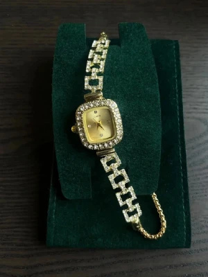 Glittrig guldfärgad klocka med strass - Snygg guldfärgad klocka med fyrkantigt ur och glittrande kristaller runt hela boetten och länken. Länken har ett justerbart band med små kulor i ändarna. Perfekt för dig som vill ha en riktigt glamorös accessoar.