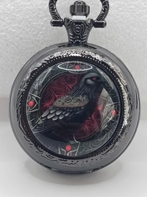 Svart fickur med korpdesign "klocka " - Unikt fickur i svart metall med detaljerad korpillustration på framsidan. Uret har en rund form och dekorerade kanter, samt röda accenter runt motivet. Perfekt för dig som gillar mystiska och alternativa accessoarer.