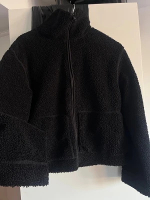 Svart teddyjacka med dragkedja - Mysig svart teddyjacka med hög krage och hel dragkedja framtill. Jackan har en boxig passform och är gjord i fluffigt, mjukt teddy-material som håller dig varm. Perfekt för lager på lager och en chill look.