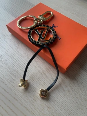  Vuitton nyckelring och accessoar - Säljer en exklusiv Vuitton nyckelring i guld och svart med LV-logga och flätad detalj. Medföljer även en svart accessoar med guldfärgade blommor i metall. Perfekt för att piffa upp väskan eller nyckelknippan.