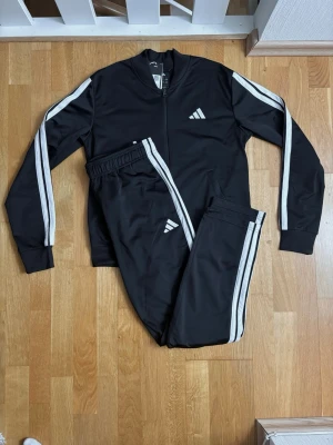 Svart Adidas träningsset dam - Svart träningsset från Adidas för dam med vita ränder längs ärmar och ben. Setet består av zip-jacka och byxor i mjukt, stretchigt material. Adidas-logga på både jacka och byxa. Perfekt för träning eller chill. Passar för dam S och herr XS. 