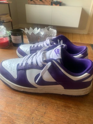 Nike Dunk Low lila och vita sneakers - Nike Dunk Low sneakers i lila och vitt med klassisk låg siluett. Ovandel i läder, lila detaljer och vit bas, samt matchande lila yttersula. Ikonisk Nike-logga på tungan och sidan. Perfekta för dig som gillar streetwear och vill sticka ut med färg. Knappt använda Max 5 gånger. Crep protect creaseskydd kommer även med som är tillför att skorna ska de så fräscha ut som möjligt och lukta gott.