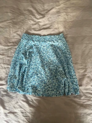 Blommig blå kortkjol  - Supersöt blå kortkjol från lager 157 med små vita blommor och elastisk midja. Kjolen har en lätt volangkant och är gjord i ett mjukt, stretchigt material som känns bekvämt. Perfekt för sommaren. 