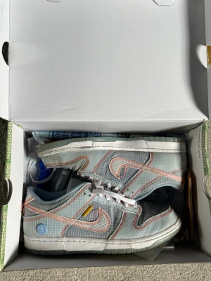 Nike Dunk Low Union LA Grey Fog - Nike Dunk Low Union LA Grey Fog sneakers med rutigt grått mesh, vita och orange detaljer samt gula och blå accenter. Swoosh-logga i orange, vit sula och unika Union-taggar på sidan. Skorna har en modern och streetinspirerad look med låg profil.