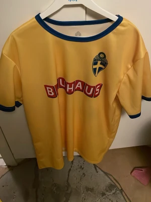 Svensk gul fotbollströja nr 22 - Gul fotbollströja från svenska landslaget med blå detaljer vid krage och ärmslut. Bauhaus-logga på bröstet och ryggen samt nummer 22 på ryggen. Perfekt för dig som vill stötta Sverige på match eller träning.